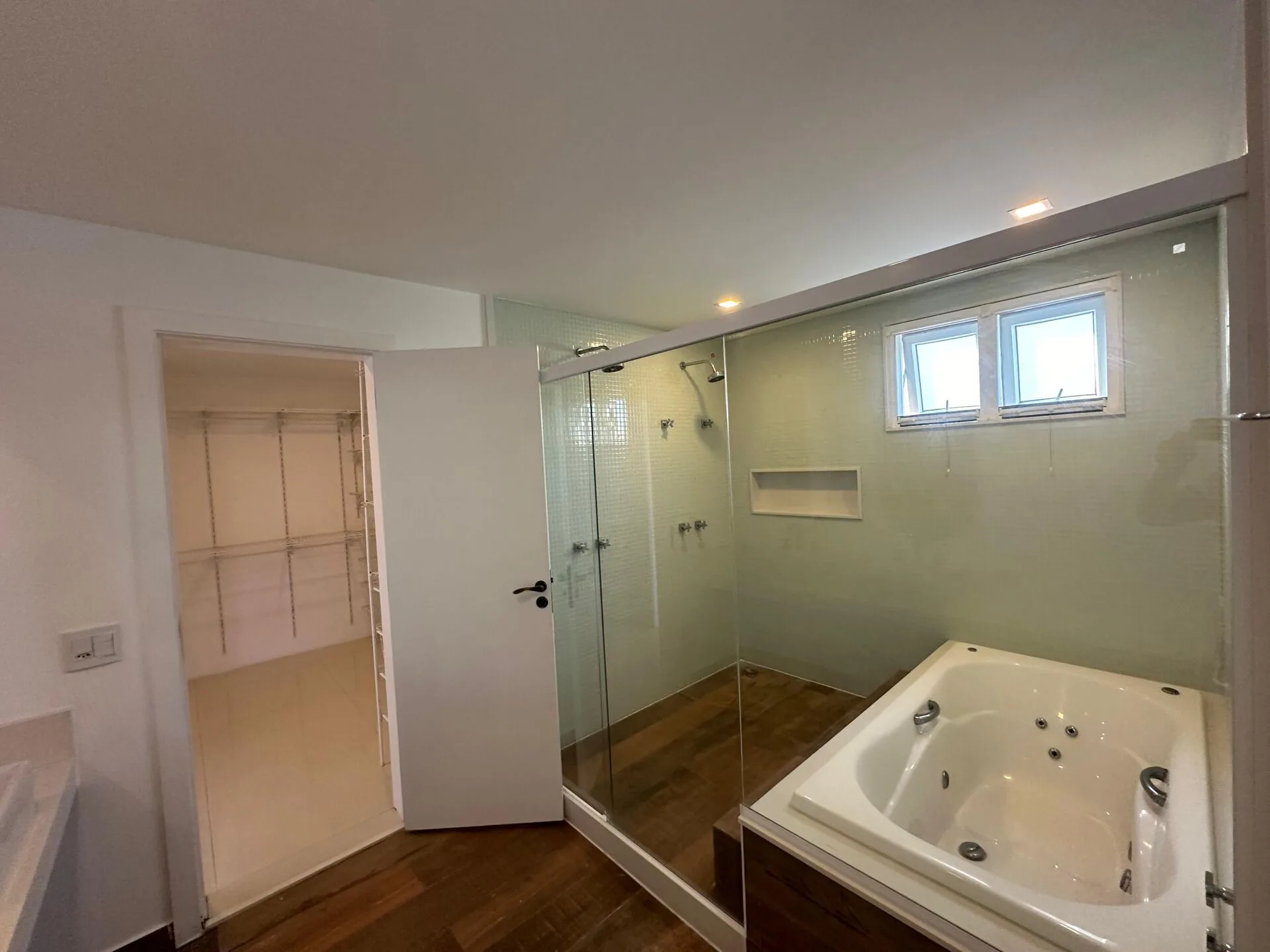 Banheiro da suíte master com banheira de hidromassagem, box de vidro com pastilhas, piso de madeira e acesso ao closet