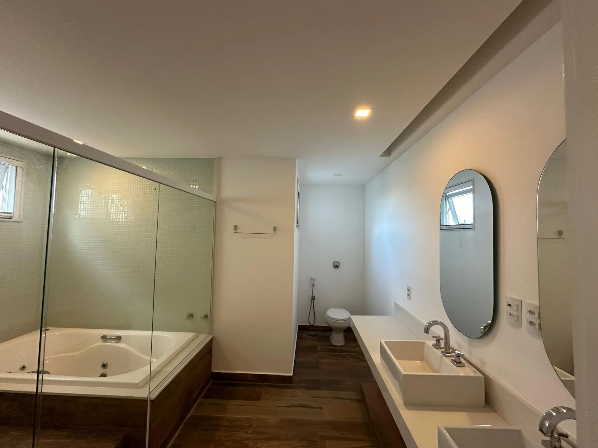 Banheiro da suíte com banheira de hidromassagem, bancada com cuba de apoio, espelho oval e box de vidro com pastilhas