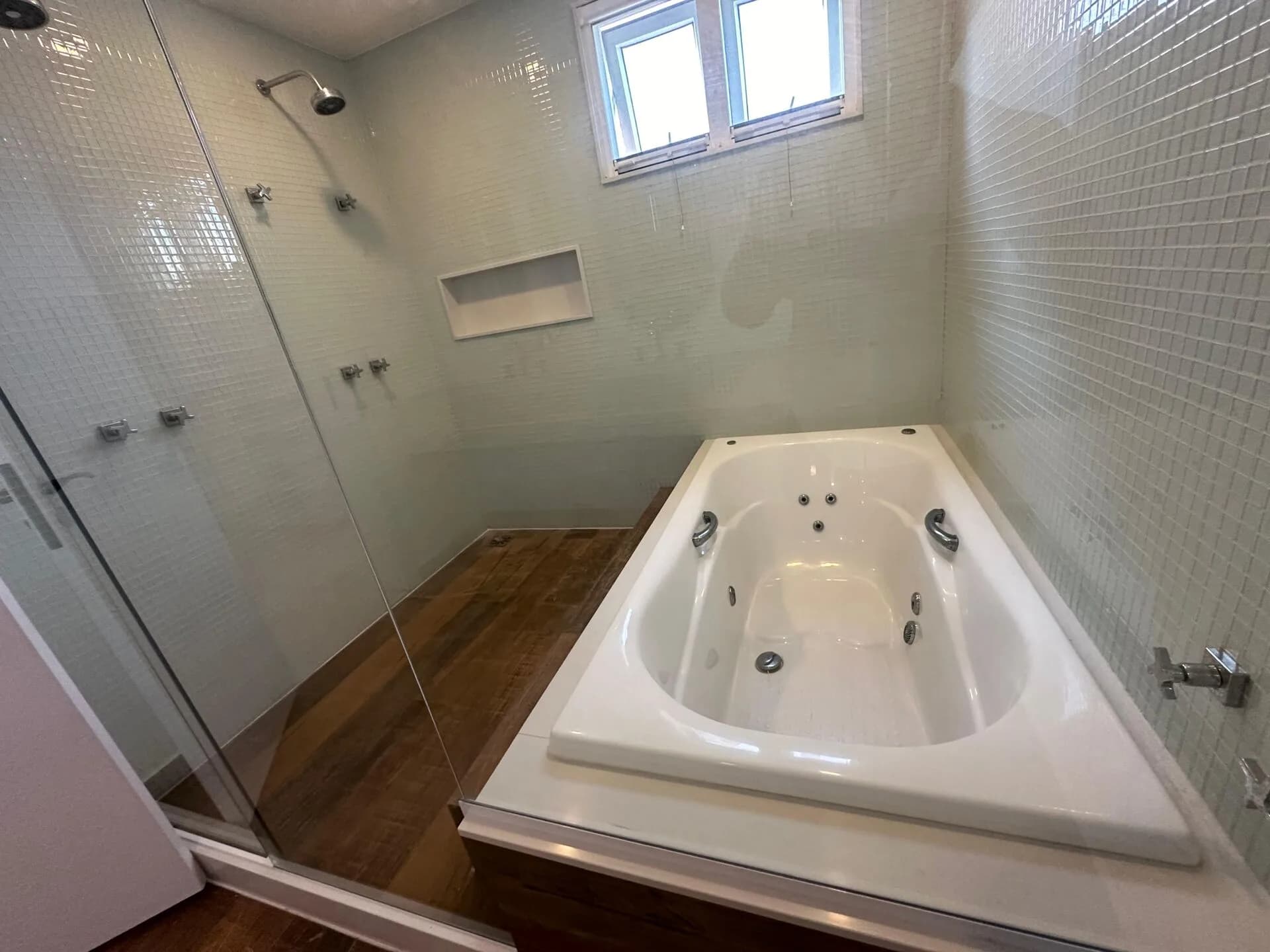 Detalhe da banheira de hidromassagem com chuveiro duplo, pastilhas verdes e piso porcelanato tipo madeira