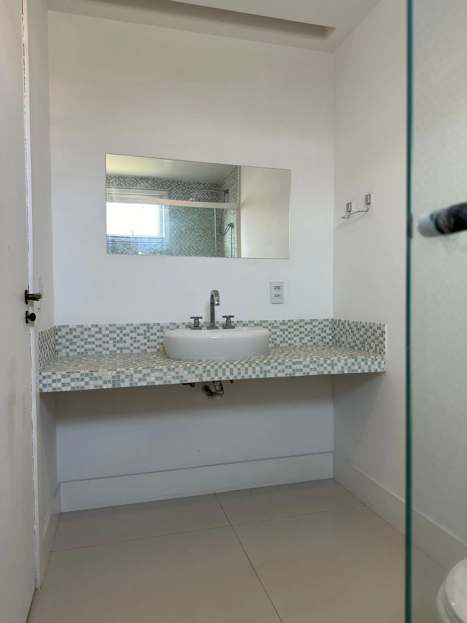 Banheiro social com bancada em pastilhas verdes e brancas, cuba de apoio, espelho retangular e box de vidro