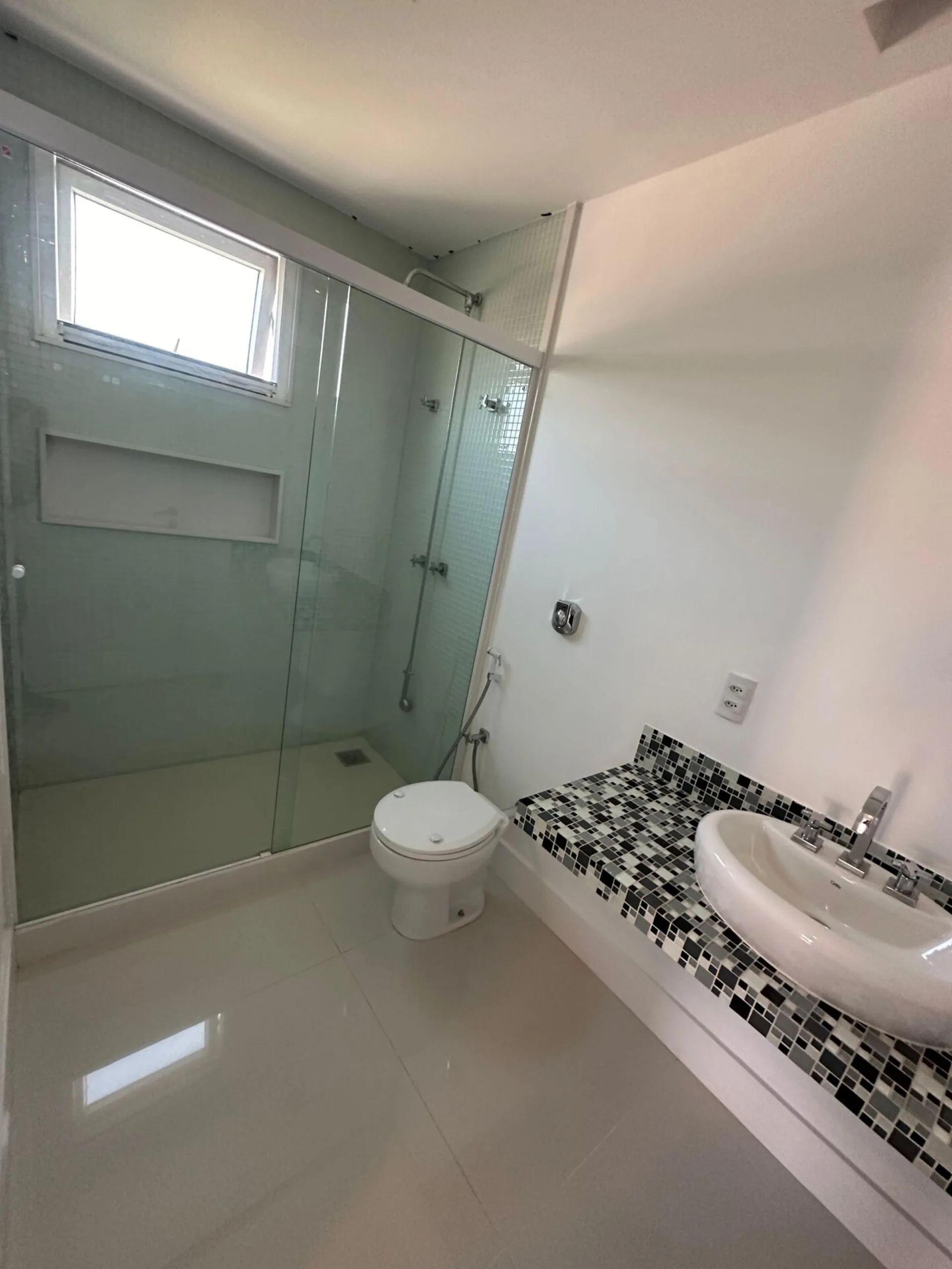 Banheiro com box de vidro, bancada em pastilhas preto e branco, cuba de apoio e piso porcelanato claro