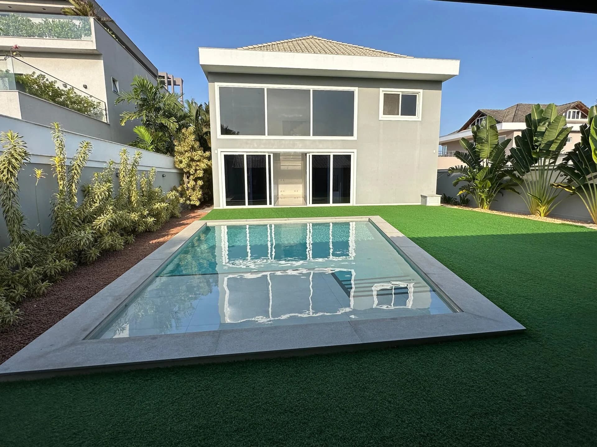 Fachada posterior da casa duplex 500m² com piscina, gramado verde e grandes janelas de vidro no Alphaville Barra da Tijuca